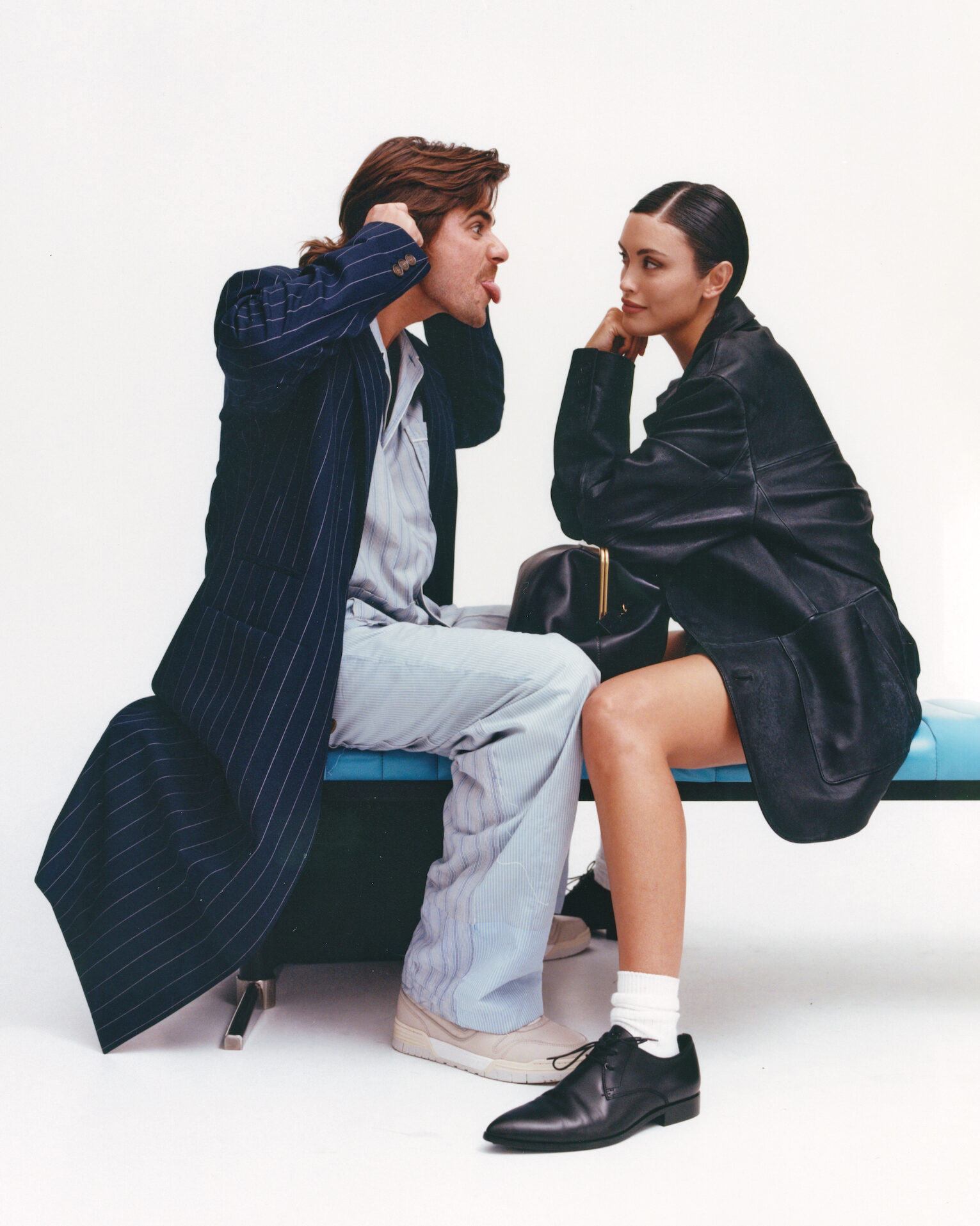 Two people pose on a turquoise bench in a white photo studio. The man is sticking his tongue out making a funny face, while the woman looks on.