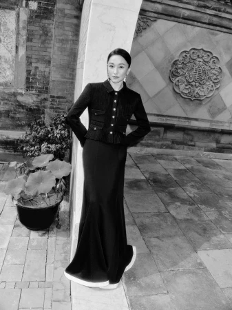 Elegant woman in a black skirt and jacket posing on a stone patio with a decorative wall, plants, and neutral color palette.