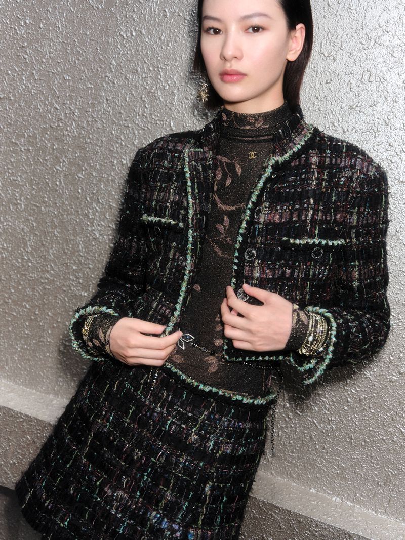 An Asian woman poses indoors wearing a black tweed skirt suit with embellishments, accessorized with statement earrings and bracelets.