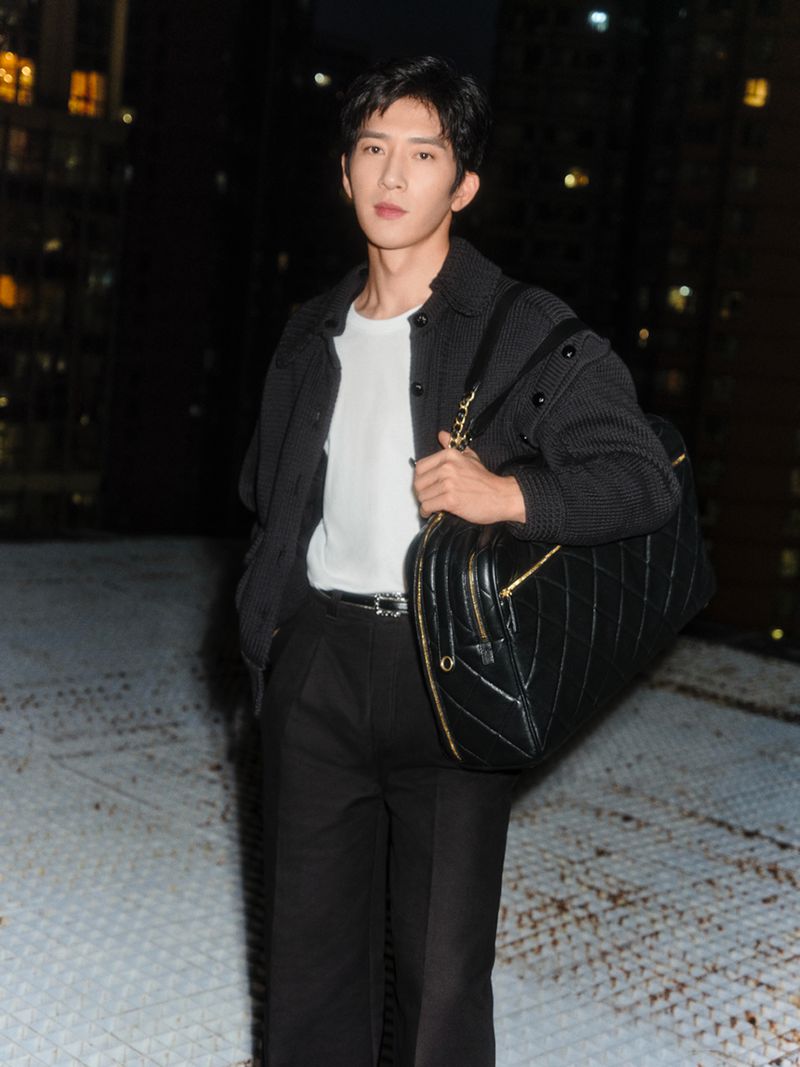 Adult male in black jacket with black leather bag stands outdoors at night with buildings in the background, fashion lifestyle shoot.