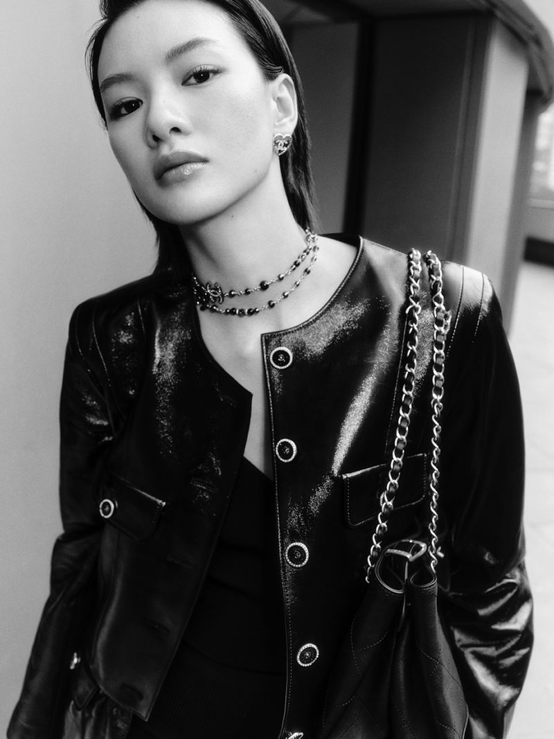 Monochrome portrait of an Asian woman with dark hair, wearing a shiny black jacket, necklace, and earrings looking at the camera.