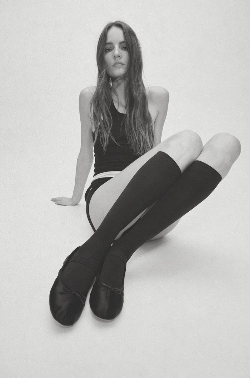 Model Tilda Lindstam seated on a white floor wearing a black tank top, knee-high socks, and black ballet flats.