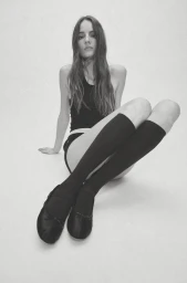Model Tilda Lindstam seated on a white floor wearing a black tank top, knee-high socks, and black ballet flats.