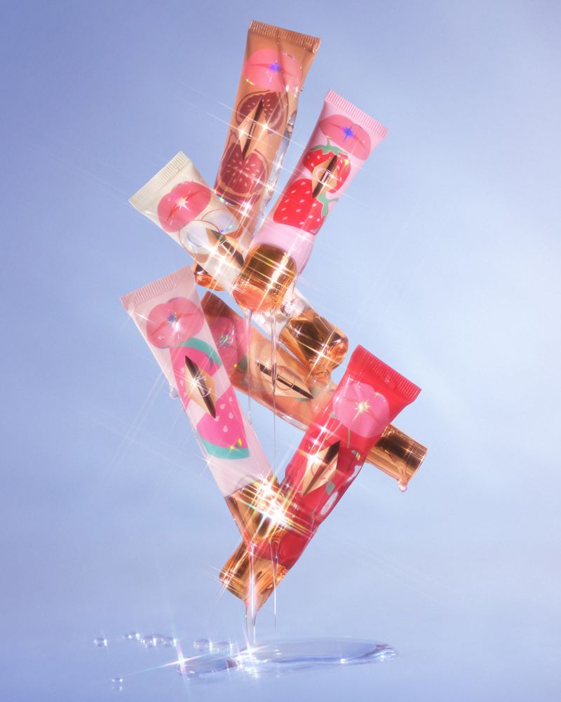 Aesthetic still life of stacked lip gloss tubes in shades of pink, red, and peach, with shimmering reflections against a blue background.