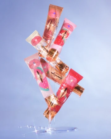 Aesthetic still life of stacked lip gloss tubes in shades of pink, red, and peach, with shimmering reflections against a blue background.