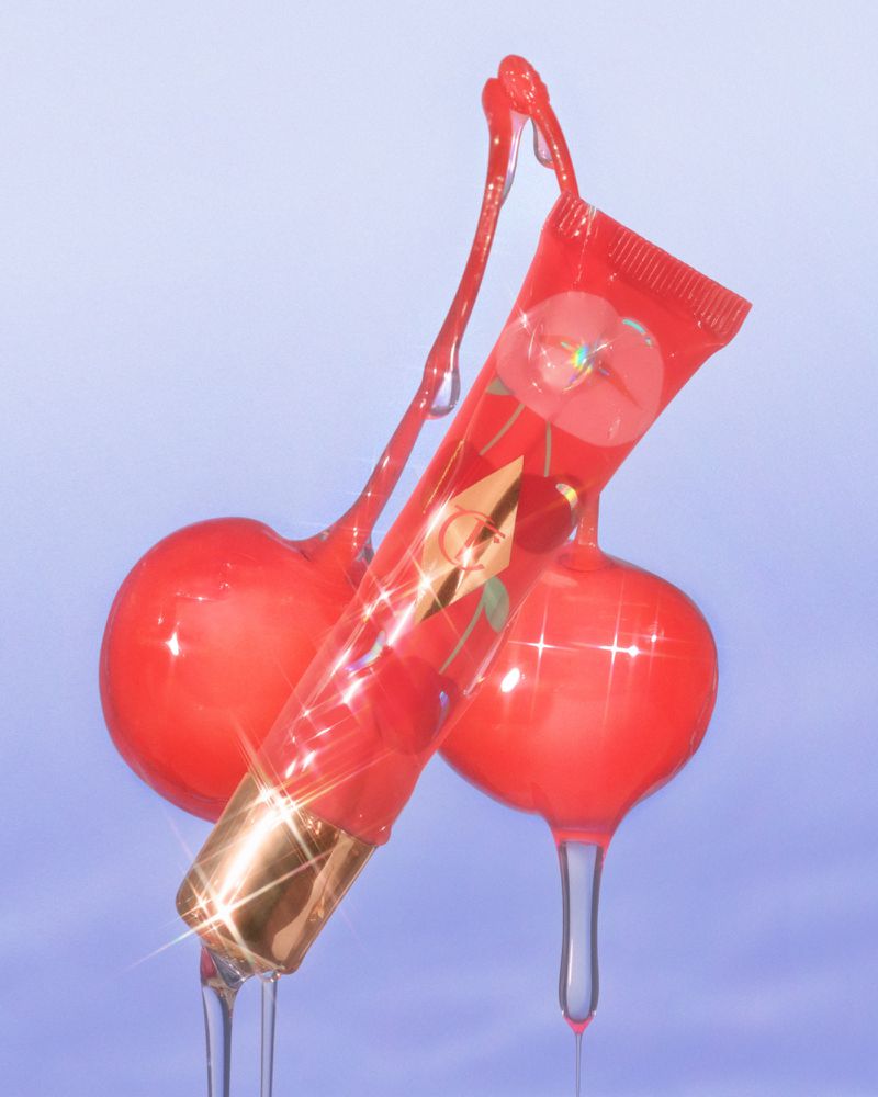 Red lip gloss in clear tube, angled with gold cap and cherry design, next to glossy cherries with clear dripping gel on blue background.