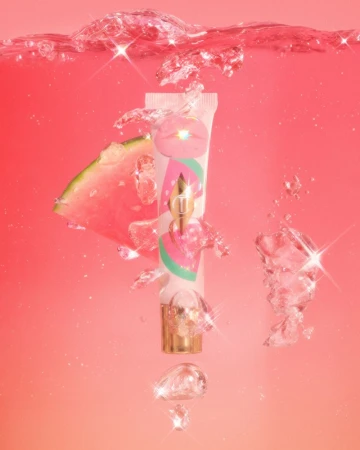 Promotional beauty shot of a tube of lip gloss with a gold applicator, watermelon slice, and water bubbles on a pink gradient background.