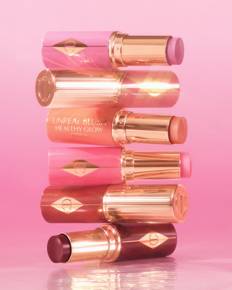 Stacked makeup sticks in shades of pink, peach, and brown with gold accents displayed on a glossy pink surface.