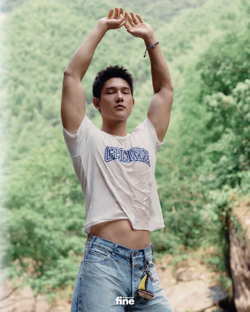 Athletic man poses with arms raised outdoors in a white tee and jeans against a background of lush green trees and a clear sky.