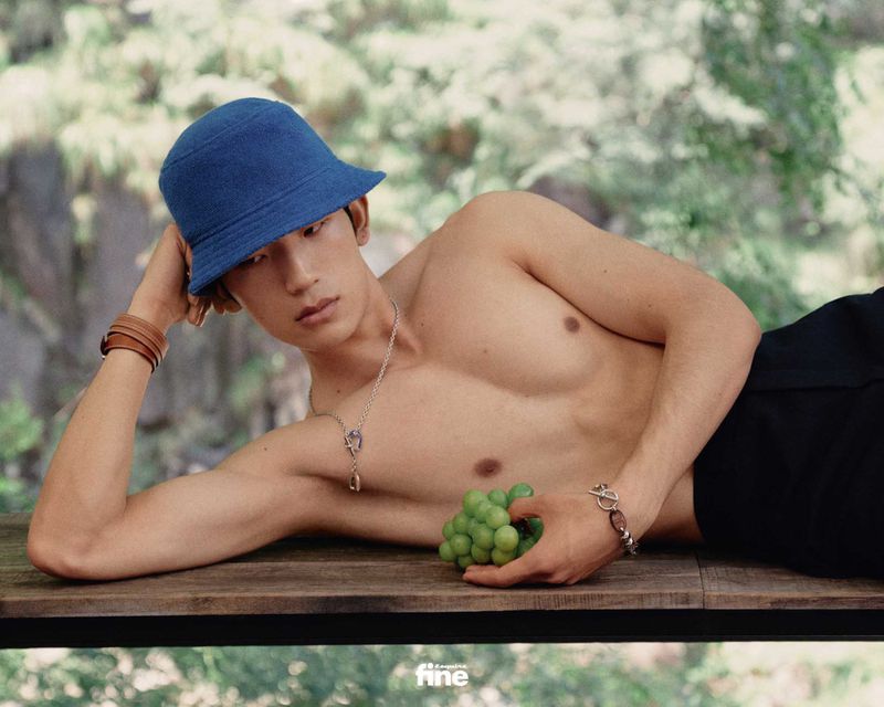 An adult male in a bucket hat poses topless with grapes on a wooden surface outdoors with a blurred background.