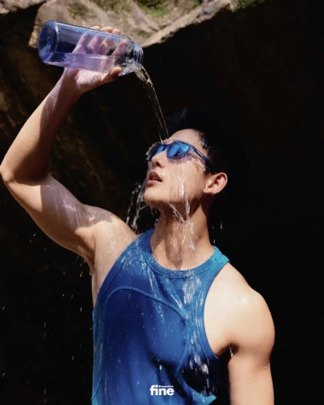 An athletic man in sunglasses pours water from a blue bottle over his head in an outdoor setting with natural rock formations.