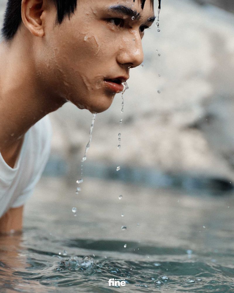 Close-up shot of a young adult male partially submerged in water with water droplets cascading from his face and hair in an outdoor setting.