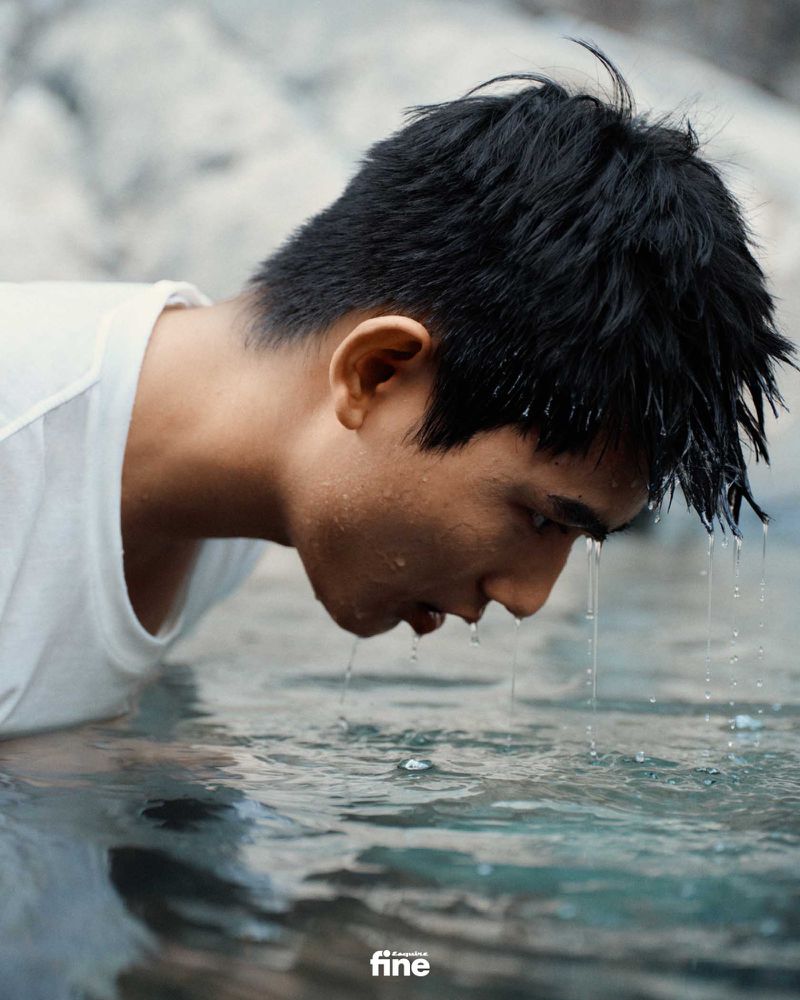 Close-up of a young Asian man drinking clear water outdoors, wearing a white T-shirt, against a background of light grey rocks and light. 