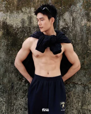 Shirtless man wearing dark sweatpants and sunglasses resting on his head, posed against a textured stone wall.
