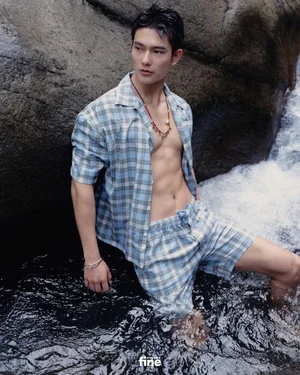 East Asian man wearing blue plaid shirt and shorts standing waist-deep in flowing water near dark rocks