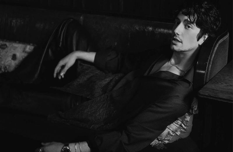 Black and white portrait of an adult male with dark, wavy hair reclining on a dark leather sofa in a dimly lit indoor environment.