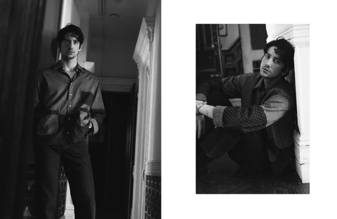 Black and white fashion portrait of a young adult male wearing a striped shirt and dark pants, posing indoors.
