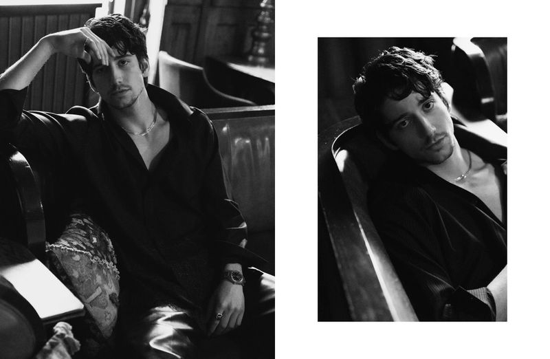 Black and white portrait of a dark-haired adult male reclining on a couch in a dark shirt and silver necklace, looking at the camera.