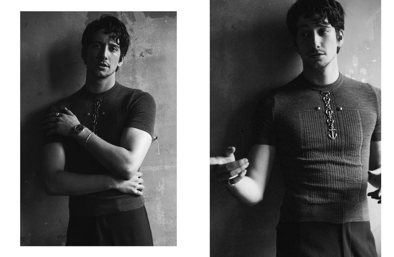 Black and white portrait of a man indoors, wearing a short-sleeved shirt with an anchor design, posing against a textured wall.