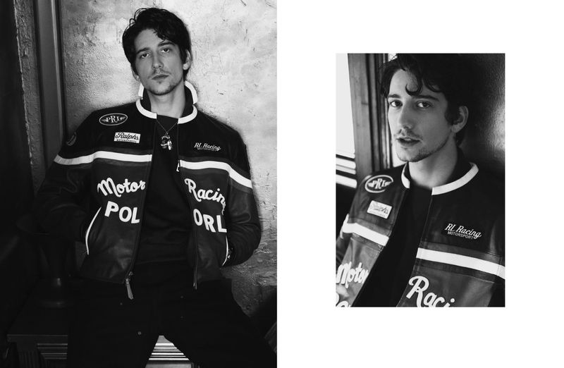 A young adult with dark hair poses in a black racing jacket with white stripes in a monochrome portrait with a neutral expression.