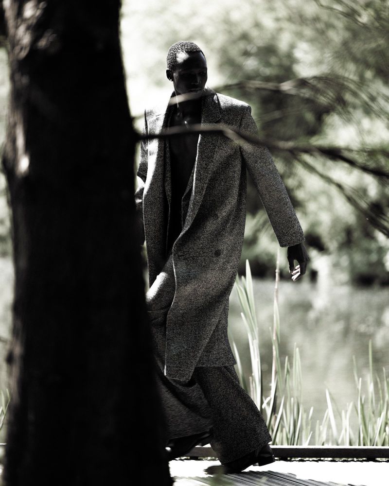 Hakim Deng walking outdoors wearing a textured long coat and wide-leg trousers behind a tree trunk in monochrome.