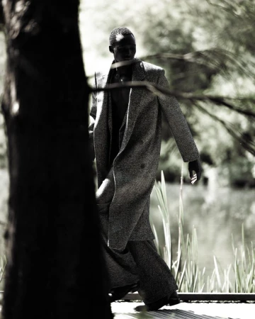 Hakim Deng walking outdoors wearing a textured long coat and wide-leg trousers behind a tree trunk in monochrome.