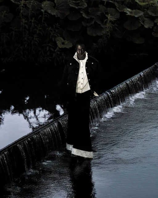 Fashion shot of a Black female model in a black jacket and skirt, standing in a river with a small waterfall, surrounded by foliage.