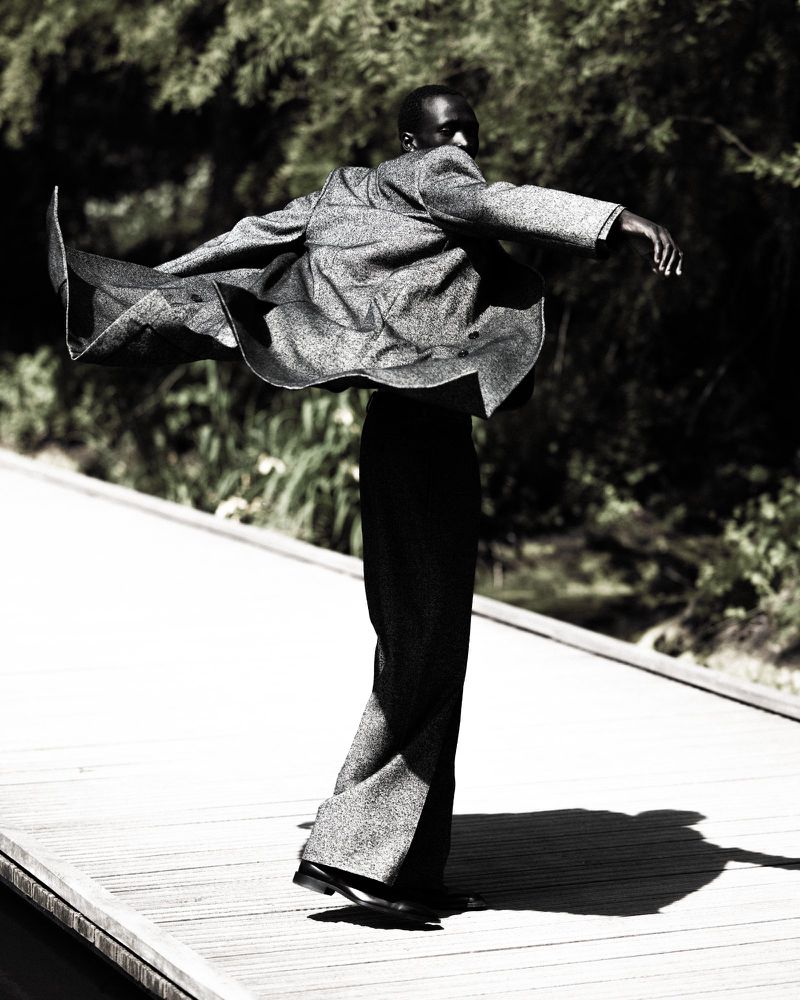 Fashion portrait of a Black male in a grey suit jacket with wide-legged pants dancing outdoors on a wooden path surrounded by trees.