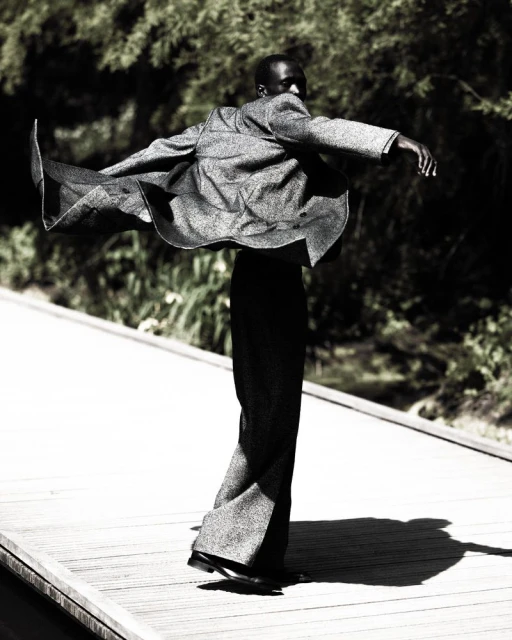 Fashion portrait of a Black male in a grey suit jacket with wide-legged pants dancing outdoors on a wooden path surrounded by trees.