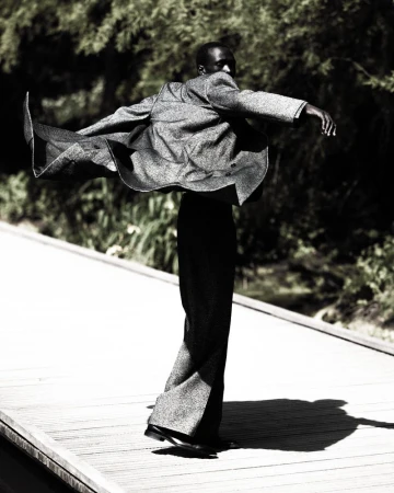 Fashion portrait of a Black male in a grey suit jacket with wide-legged pants dancing outdoors on a wooden path surrounded by trees.