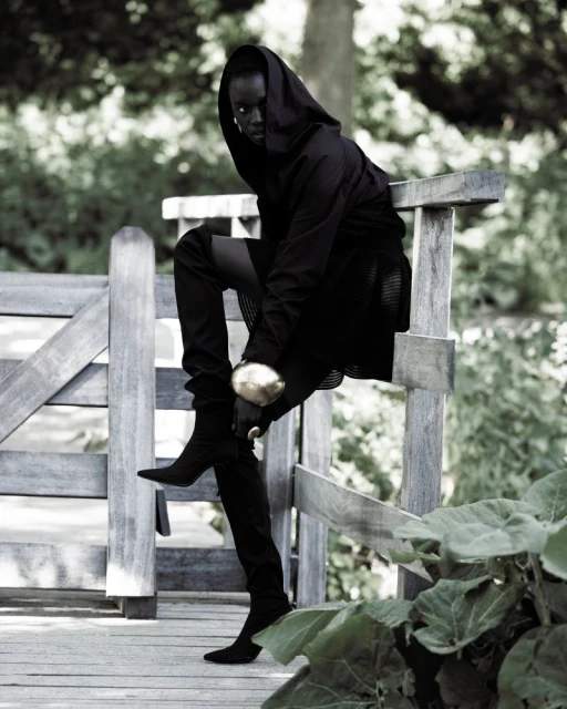 An adult female model sits on a wooden fence wearing a hooded outfit with thigh-high boots in a garden setting with lush greenery.