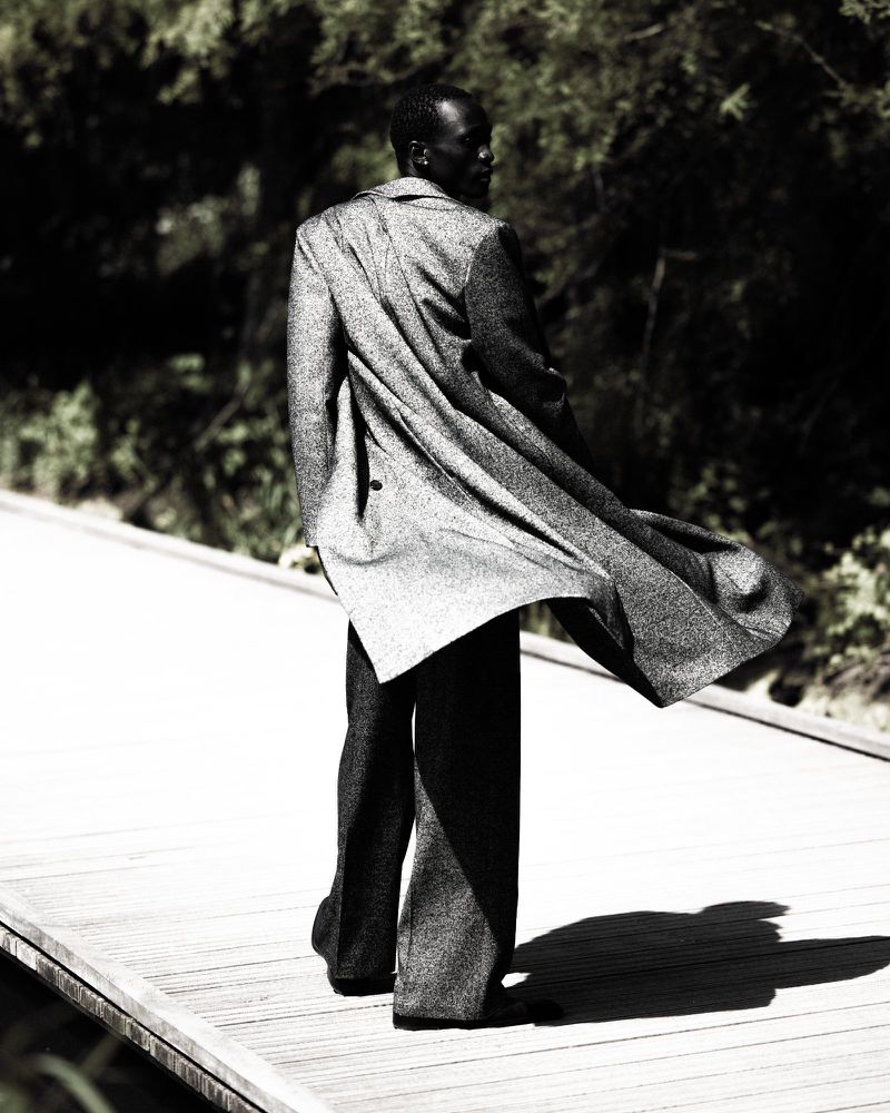 An African male fashion model in a tweed overcoat and trousers walks on a boardwalk with greenery in the background looking over his shoulder.