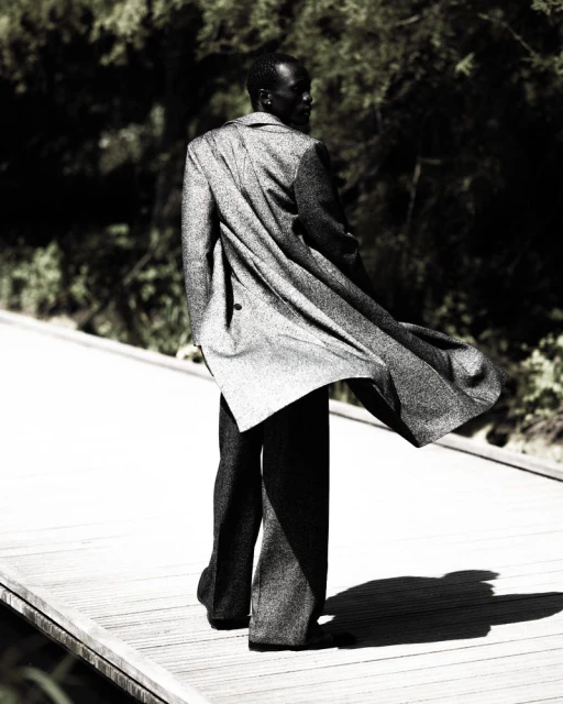 An African male fashion model in a tweed overcoat and trousers walks on a boardwalk with greenery in the background looking over his shoulder.