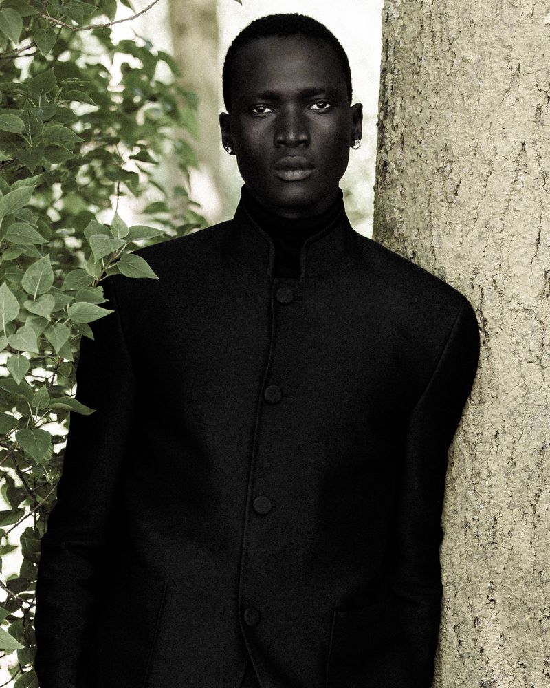 Model Hakim Deng in a black mandarin collar jacket leaning against a tree trunk beside green foliage.