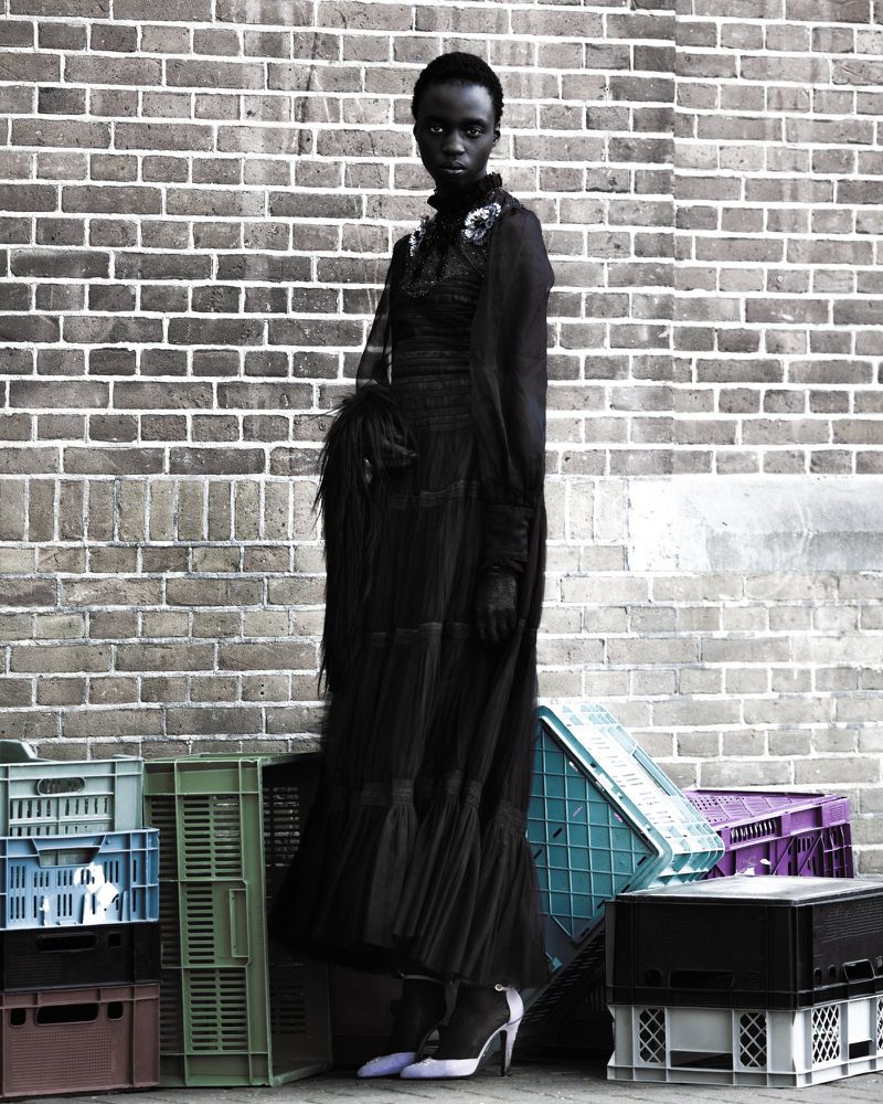 A tall, slim Black woman in a full black gown poses against a brick wall, standing in front of stacked colorful plastic crates.