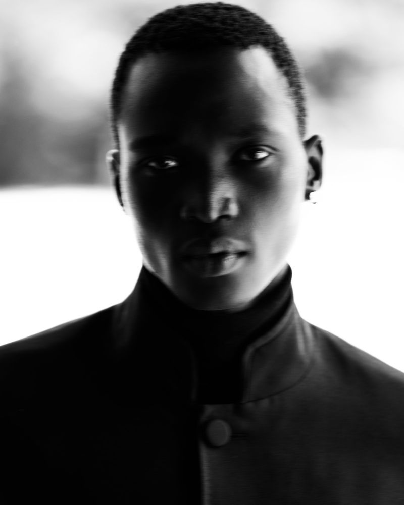 Close-up portrait of a Black male model with short hair wearing a dark jacket against an out-of-focus background in black and white.