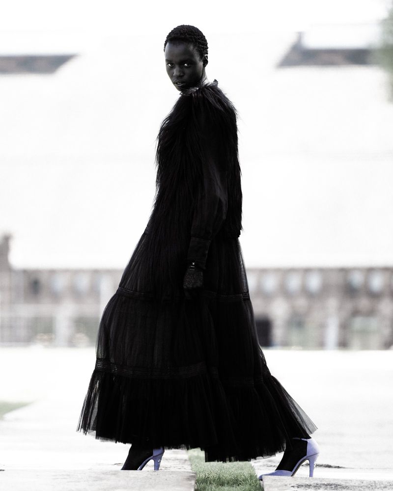 Fashion shot of a Black female model wearing a long black dress and lilac boots, walking on a concrete surface with a blurred background.