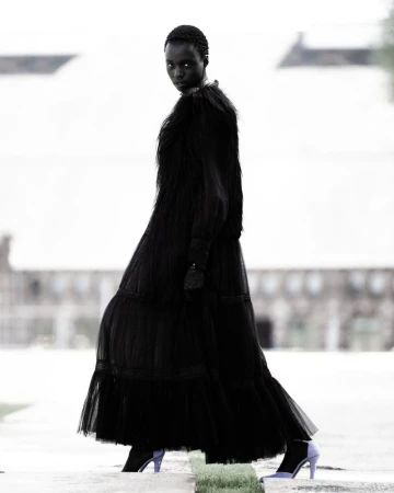 Fashion shot of a Black female model wearing a long black dress and lilac boots, walking on a concrete surface with a blurred background.
