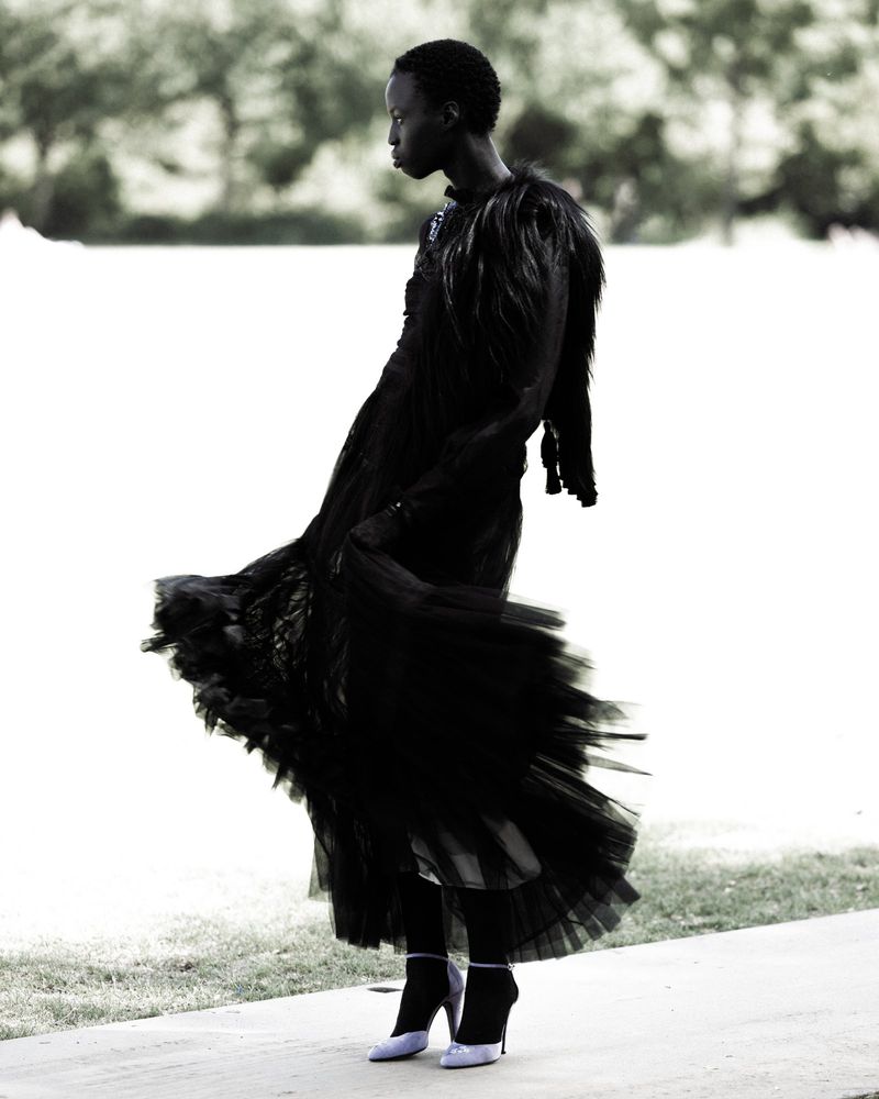 Elegant woman in a flowing black gown with feathered accents gracefully poses against a blurred natural backdrop, showcasing fashion and style.