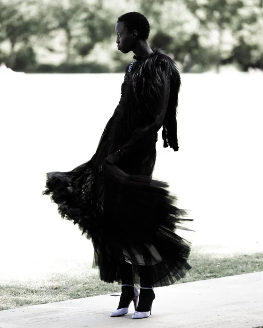 Elegant woman in a flowing black gown with feathered accents gracefully poses against a blurred natural backdrop, showcasing fashion and style.