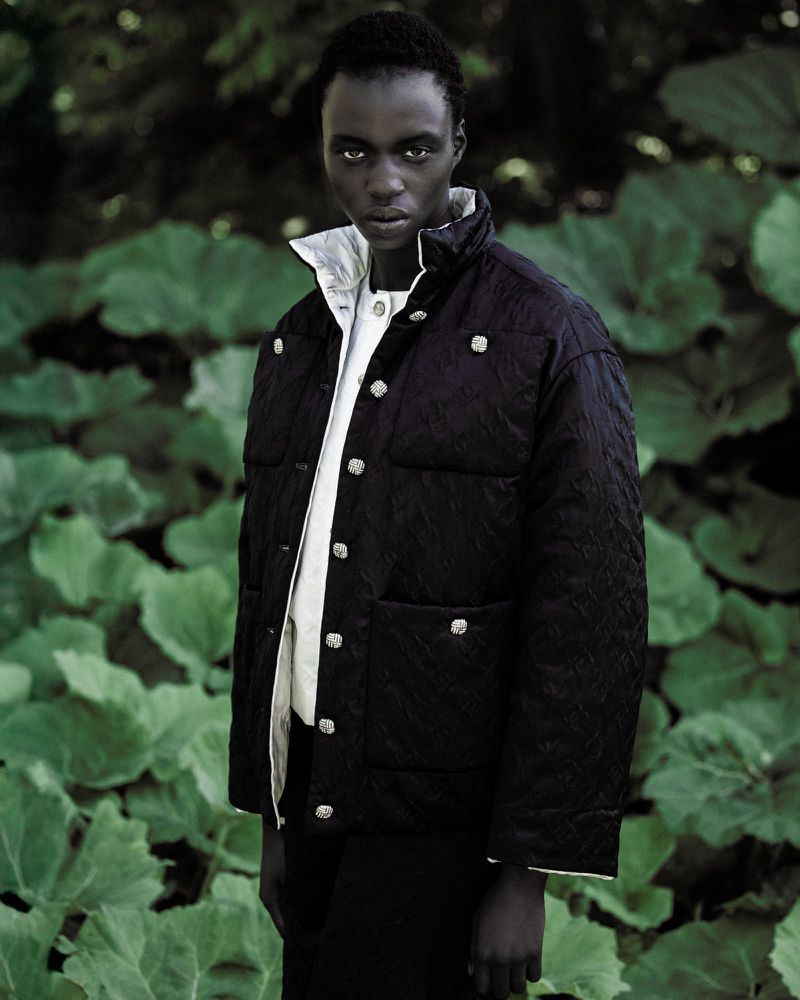 Fashion shot of a black male model wearing a quilted black jacket with silver buttons in front of a leafy green background outdoors.