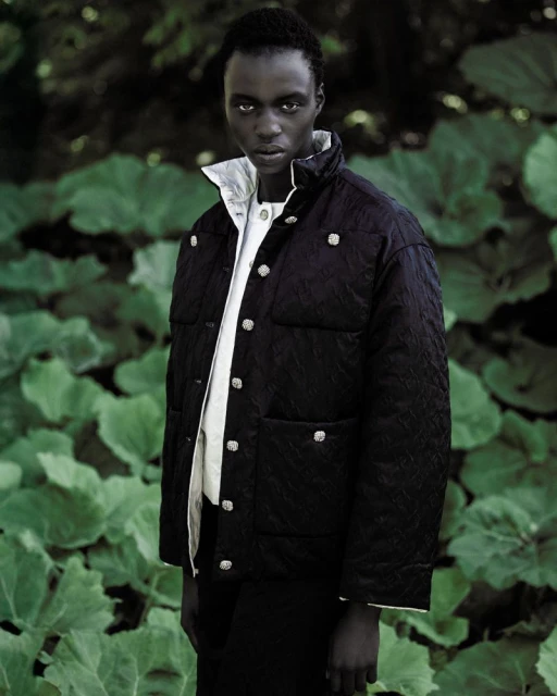 Fashion shot of a black male model wearing a quilted black jacket with silver buttons in front of a leafy green background outdoors.
