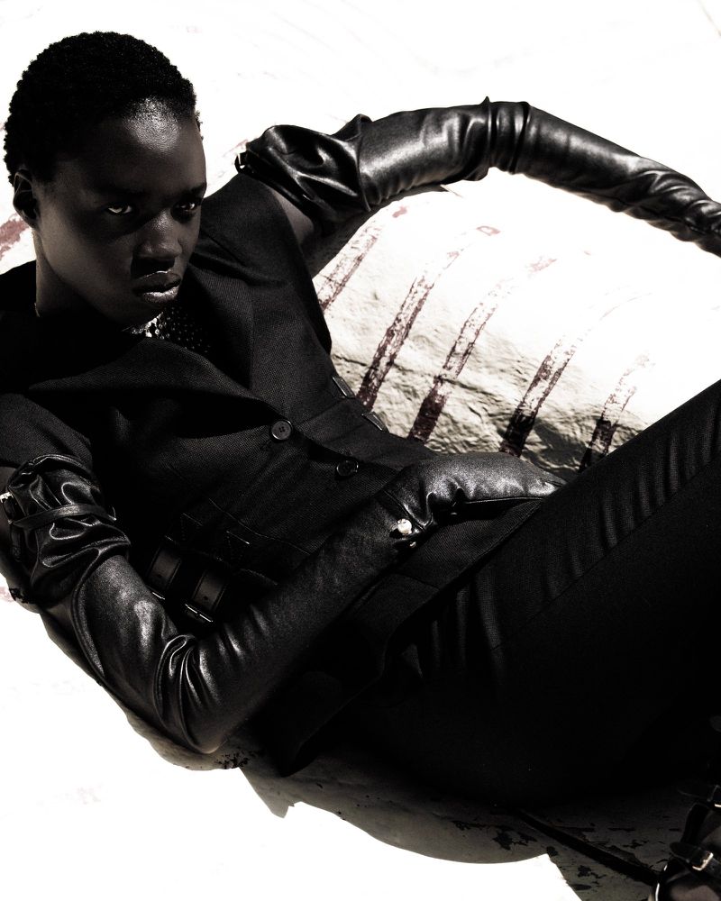 Fashion portrait of a striking Black woman wearing a black leather suit and gloves, captured in a bright studio setting with striped backdrop.