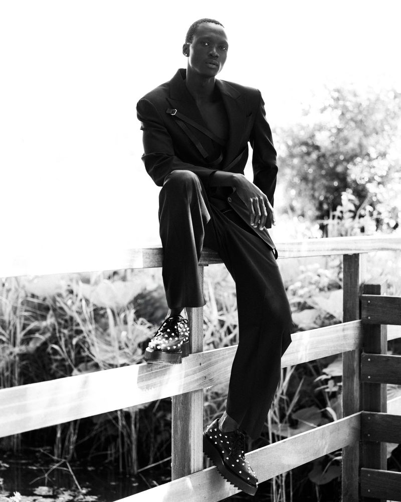A black-and-white portrait shows a slender black male model in a dark suit sitting on a wooden fence outdoors with dense plants behind him.