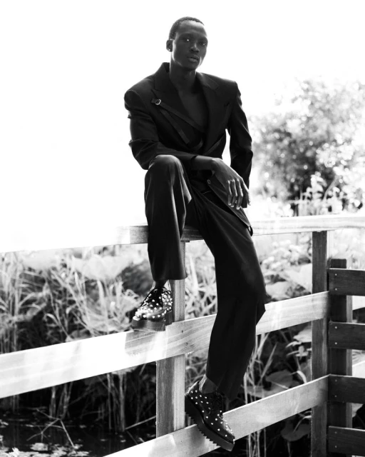 A black-and-white portrait shows a slender black male model in a dark suit sitting on a wooden fence outdoors with dense plants behind him.