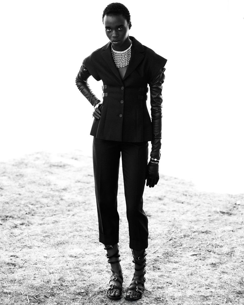 An African female model in a black jacket, pants, and gladiator sandals poses in a studio environment with a bright background.