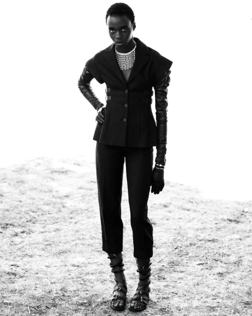 An African female model in a black jacket, pants, and gladiator sandals poses in a studio environment with a bright background.