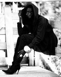 Model wearing a black hooded top and dark trousers sitting outdoors against a wooden fence structure in a monochrome fashion editorial portrait.