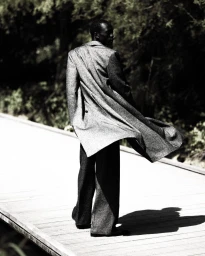Model Hakim Deng walks along a wooden boardwalk wearing a flowing tailored grey coat and matching wide-leg trousers in a park.