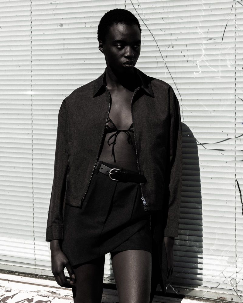 Model wearing a dark unzipped jacket over a black bra top and skirt against a ribbed shutter backdrop for Numero Netherlands.